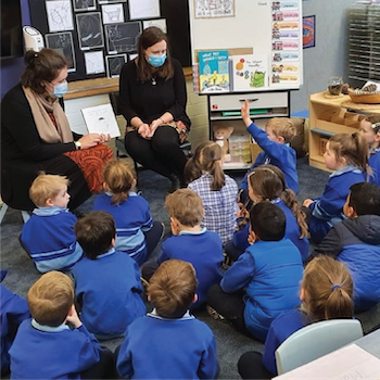 Two adults reading to a group of primary school children seated on a classroom floor.